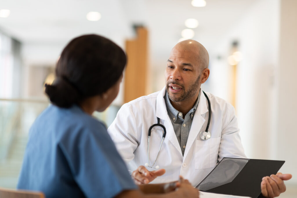 Two medical professionals sit in a hospital hallway discussing their patient's treatment. One of the doctors is holding a folder containing medical records and test results. They are consulting the data.