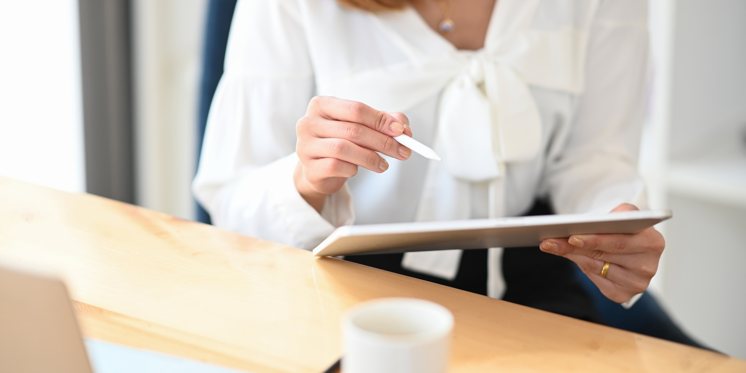 Photo of a girl holding/using a stylus pen for drawing/planning to computer tablet while sitting at the wooden working desk over comfortable living room as background.